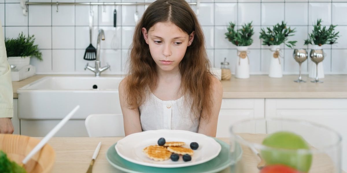 A young girl looks down unhappily at her breakfast pancakes with blueberries in a modern kitchen.