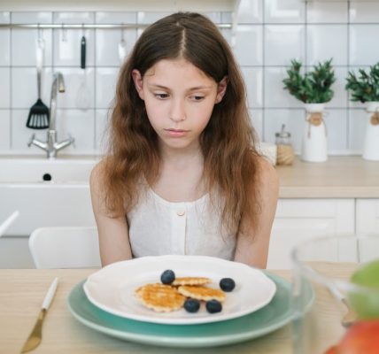 A young girl looks down unhappily at her breakfast pancakes with blueberries in a modern kitchen.