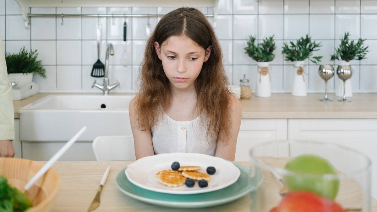 A young girl looks down unhappily at her breakfast pancakes with blueberries in a modern kitchen.